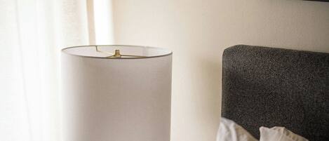 A neatly made bed with a white pillow, a bedside table with a lamp, a phone, and a power strip.