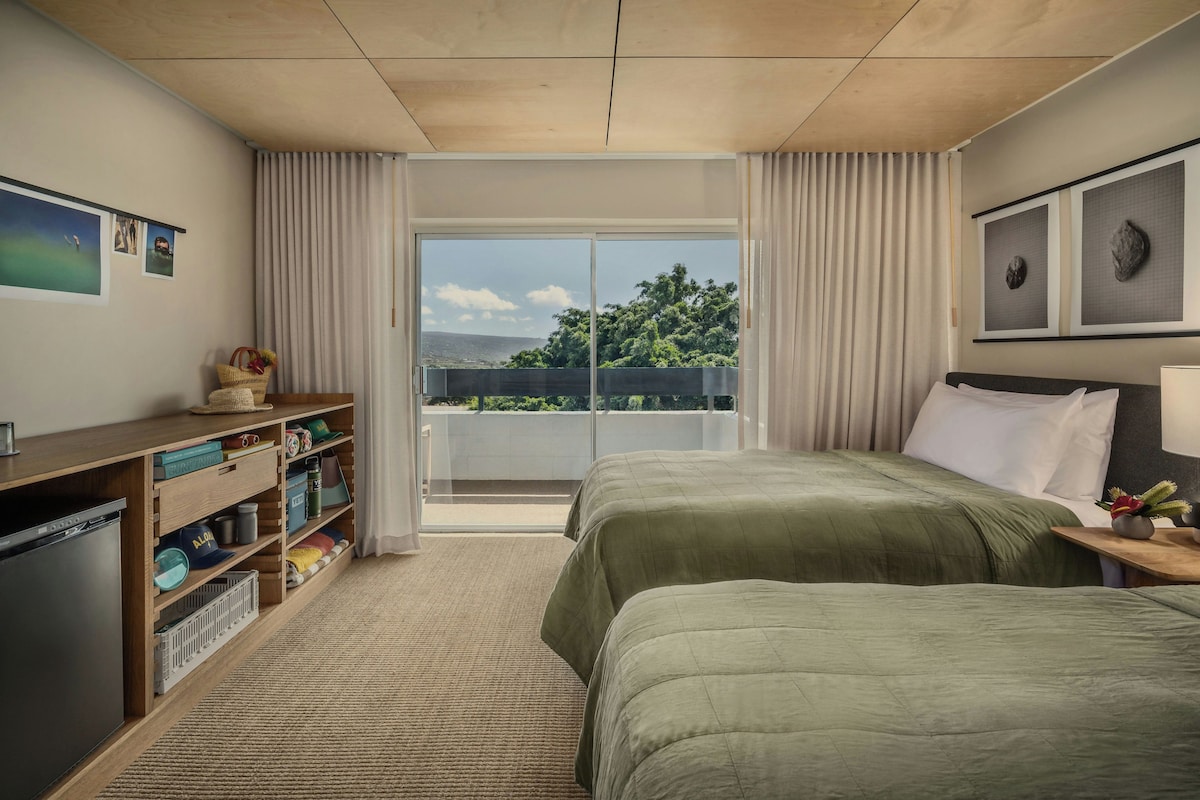 A hotel room with a bed, a desk, a bookshelf, a window with curtains, and a view of the outdoors.