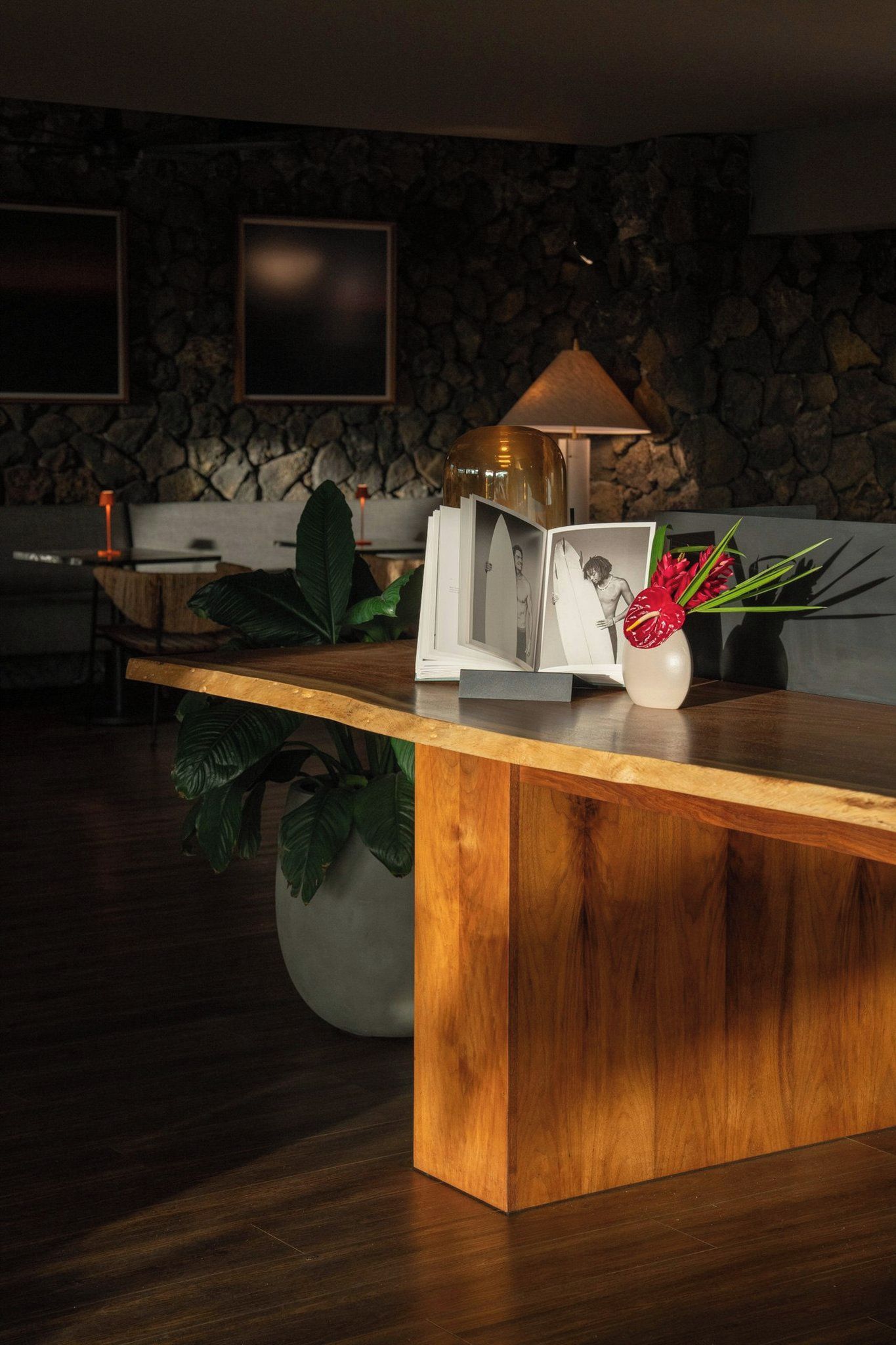 Una reception in legno con una fotografia incorniciata, un vaso con fiori rossi e una lampada, in un ambiente moderno con pareti in pietra e una pianta.