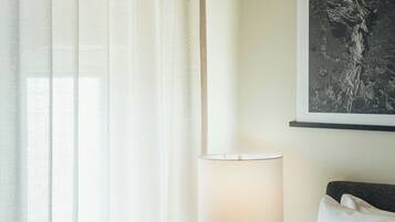 A hotel room with a bed, a bedside table with a lamp, a framed artwork on the wall, and sheer curtains.