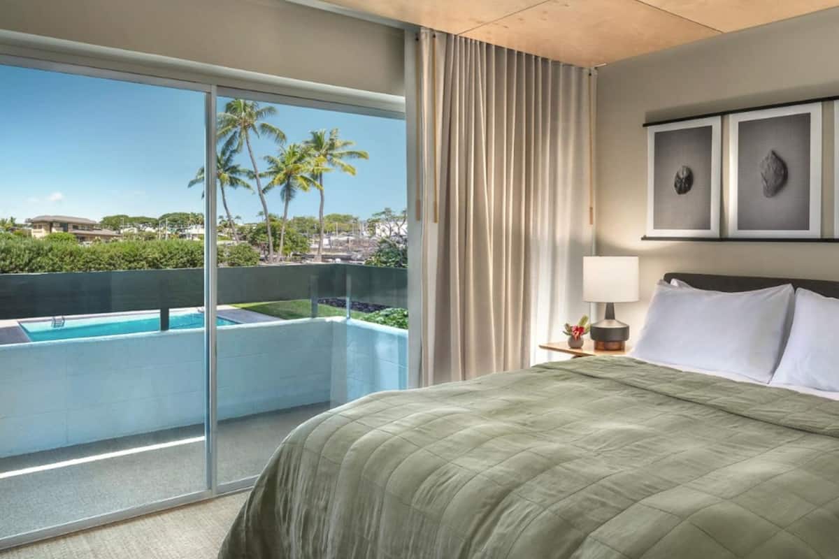 Een hotelkamer met een groot bed, een nachtkastje, een lamp en uitzicht op een zwembad en palmbomen.