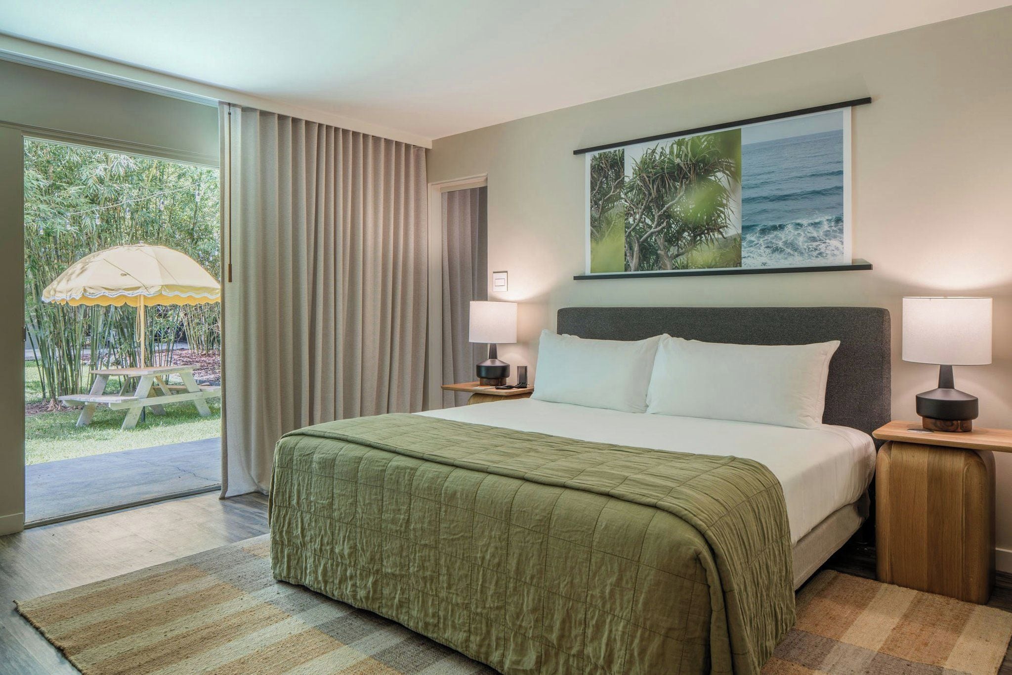 Habitación de hotel con cama, dos mesitas de noche, ventana con cortinas y vista a áreas verdes, además de un cuadro en la pared.