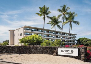 Un hôtel à plusieurs étages, avec un mur de pierre à l’avant, des palmiers et une enseigne indiquant « PACIFIC 19 ».