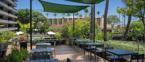 Un espace extérieur aménagé en terrasse, avec des tables et des chaises, entouré de verdure et d’une piscine.