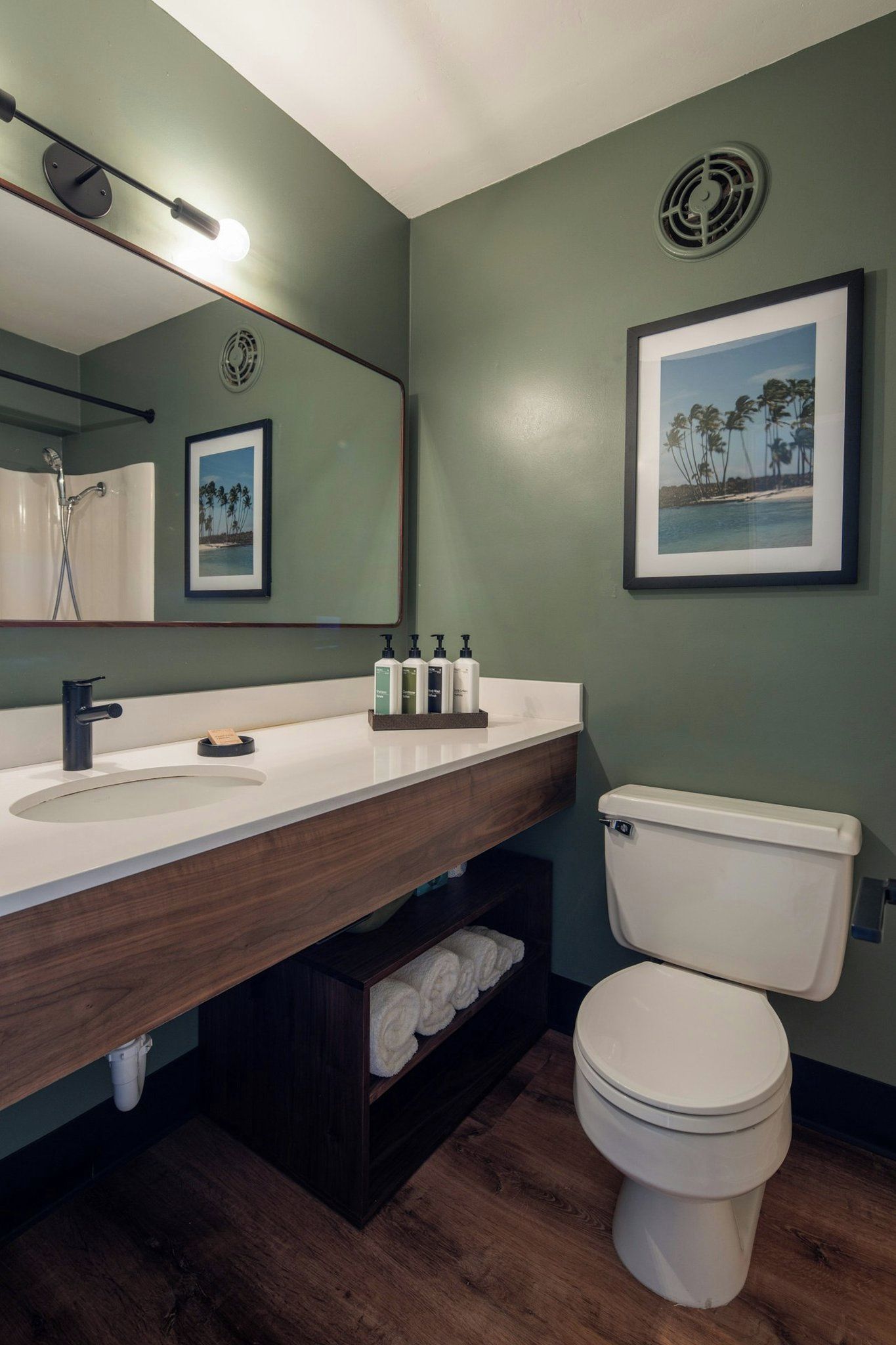 A bathroom with a wooden vanity, a white toilet, a framed picture of a beach, and a mirror with a light fixture.