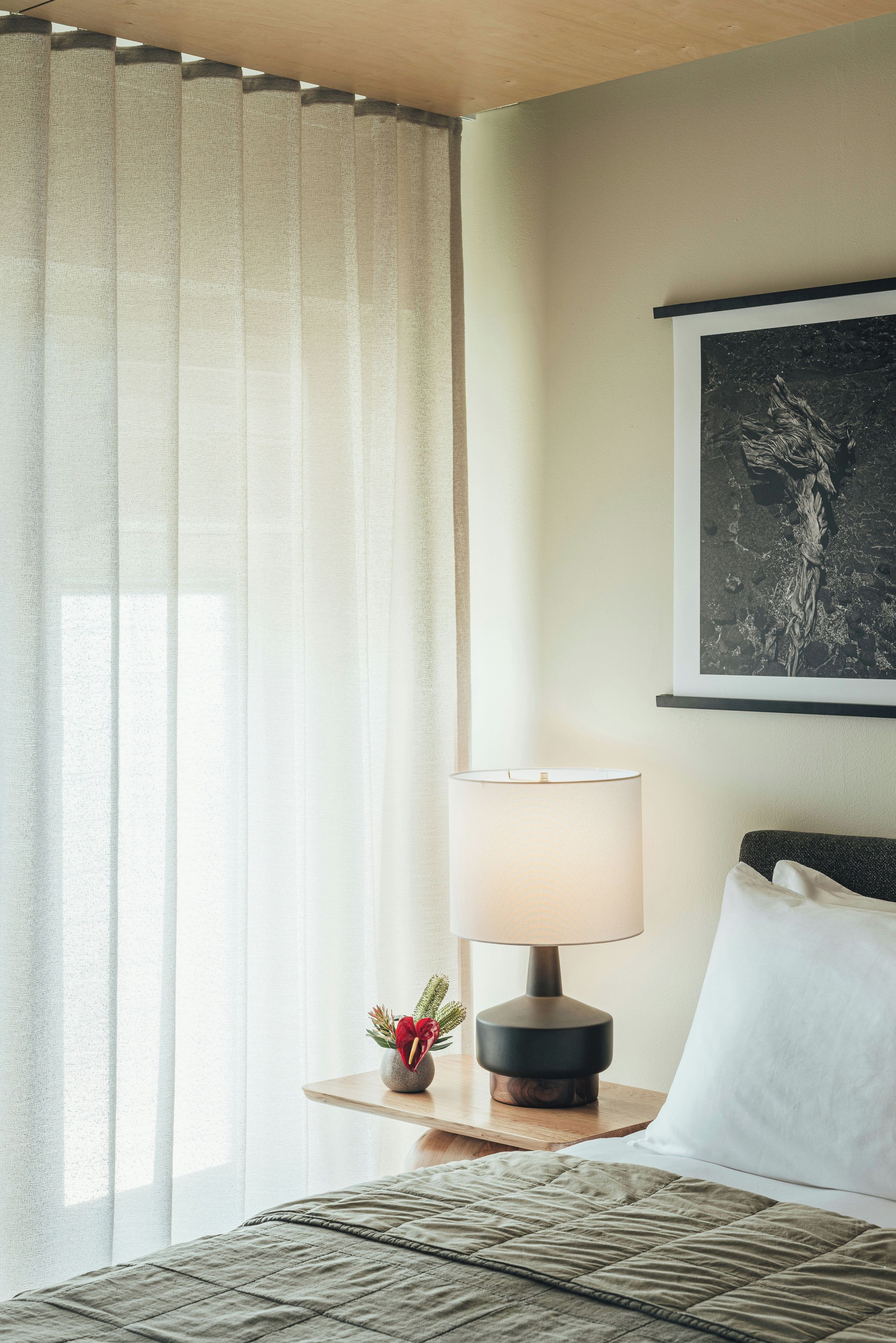 A hotel room with a bed, a bedside table with a lamp, a framed artwork on the wall, and sheer curtains.