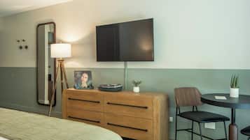 Habitación de hotel con cama, cómoda de madera, escritorio con dos sillas, televisor de pantalla plana, lámpara y una pequeña planta en maceta.