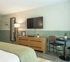 Ein Hotelzimmer mit einem Bett, einer Holzkommode, einem Schreibtisch mit zwei Stühlen, einem Flachbildfernseher, einer Lampe und einem kleinen Topfpflanzen.