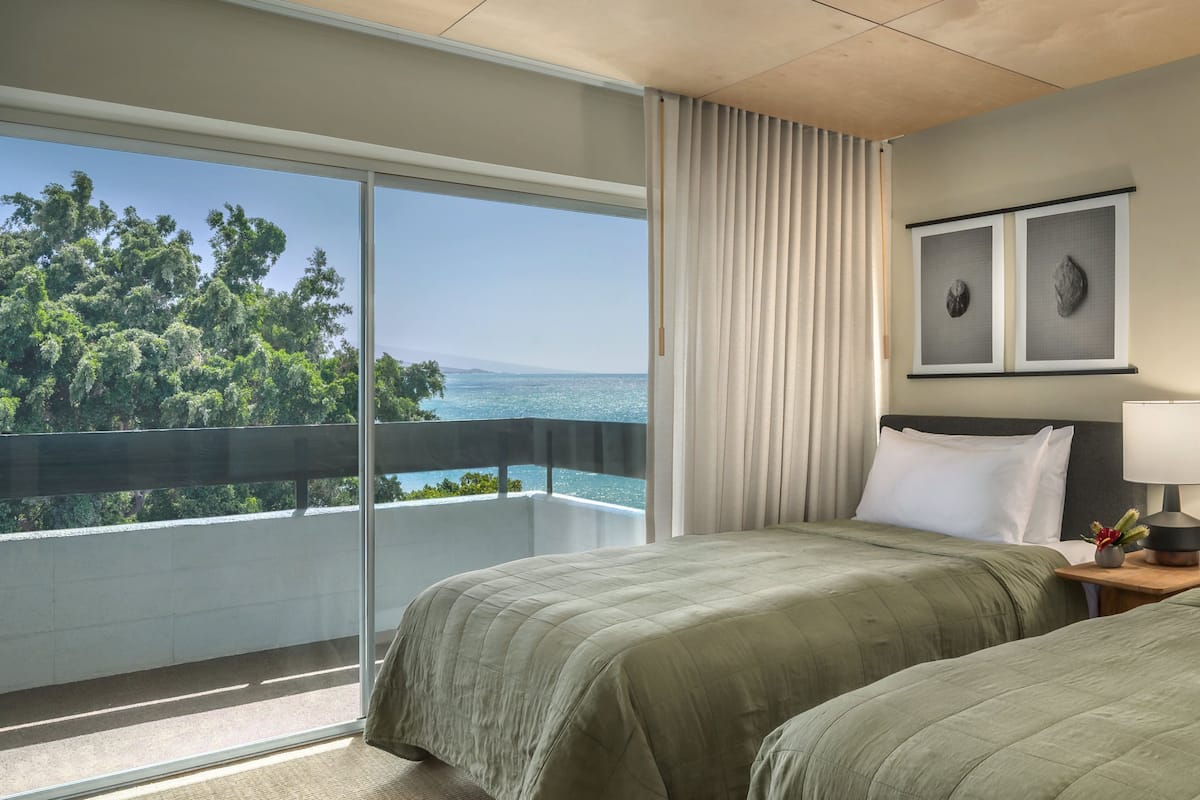 Een hotelkamer met twee bedden, een balkon met uitzicht op bomen en de zee, en kunstwerken in lijsten aan de muur.