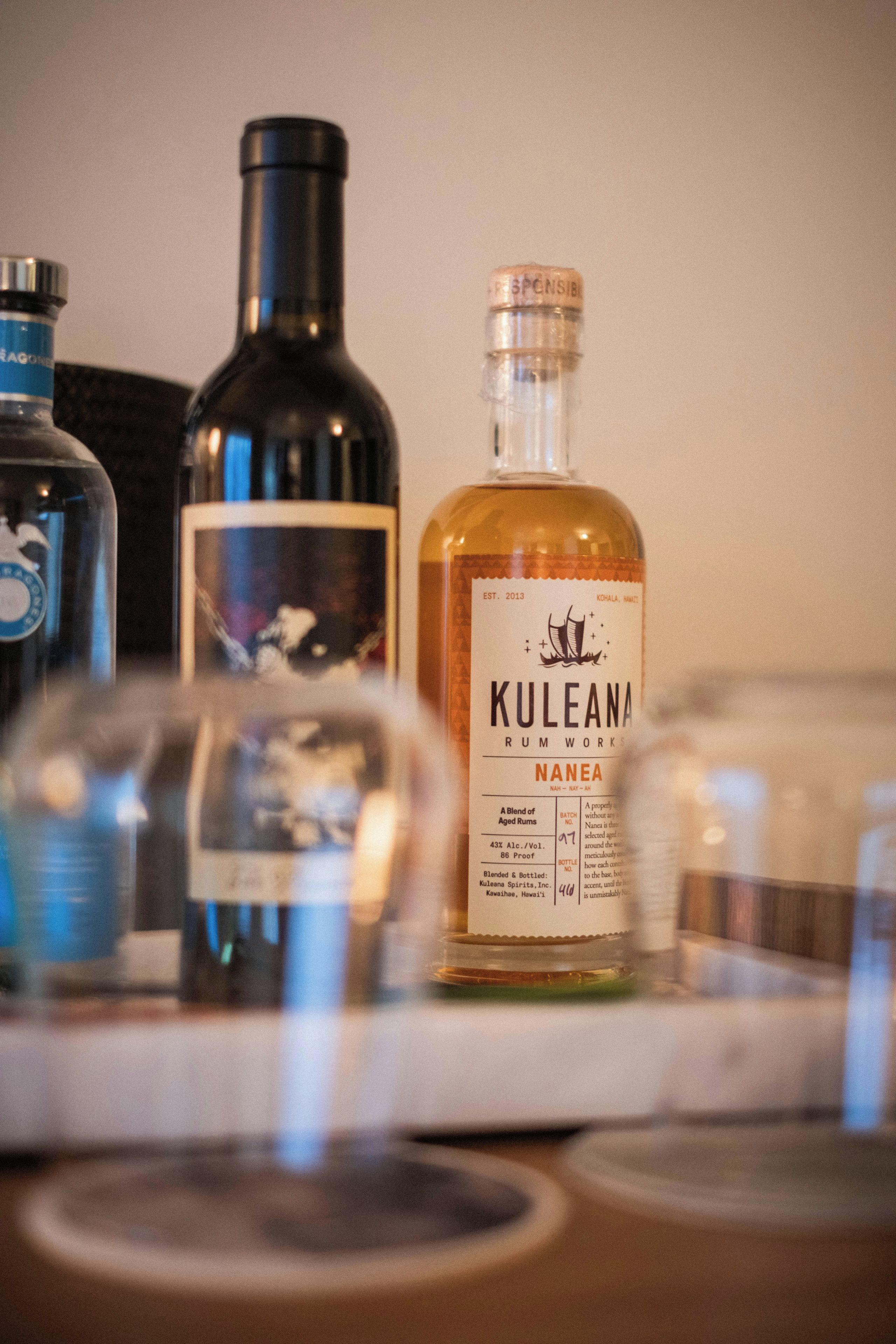 Una botella de ron Kuleana con etiqueta, una botella de otra bebida espirituosa y un vaso en primer plano.