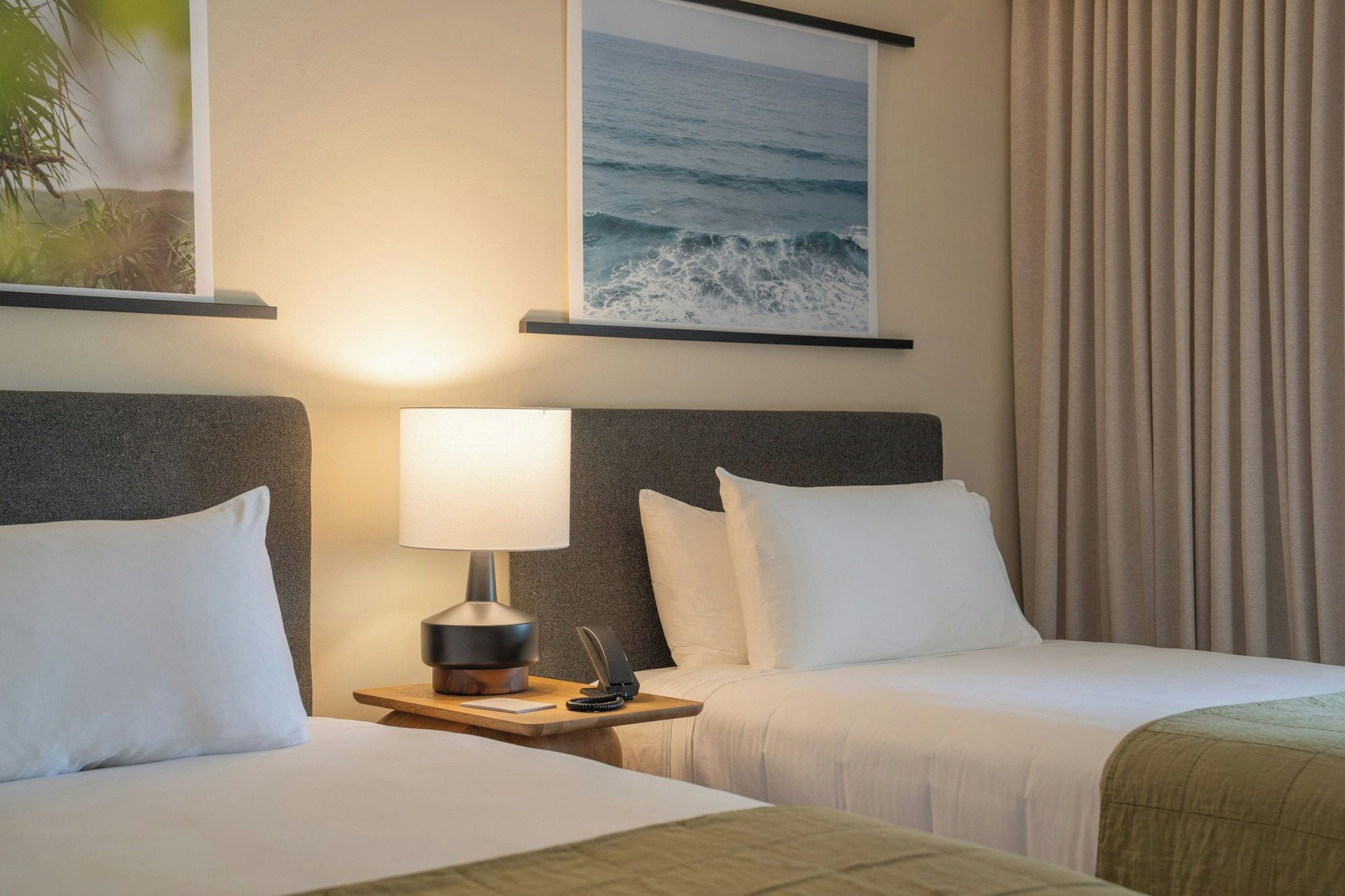 Habitación de hotel con dos camas, una mesita de noche con lámpara, teléfono y un cuadro del océano.