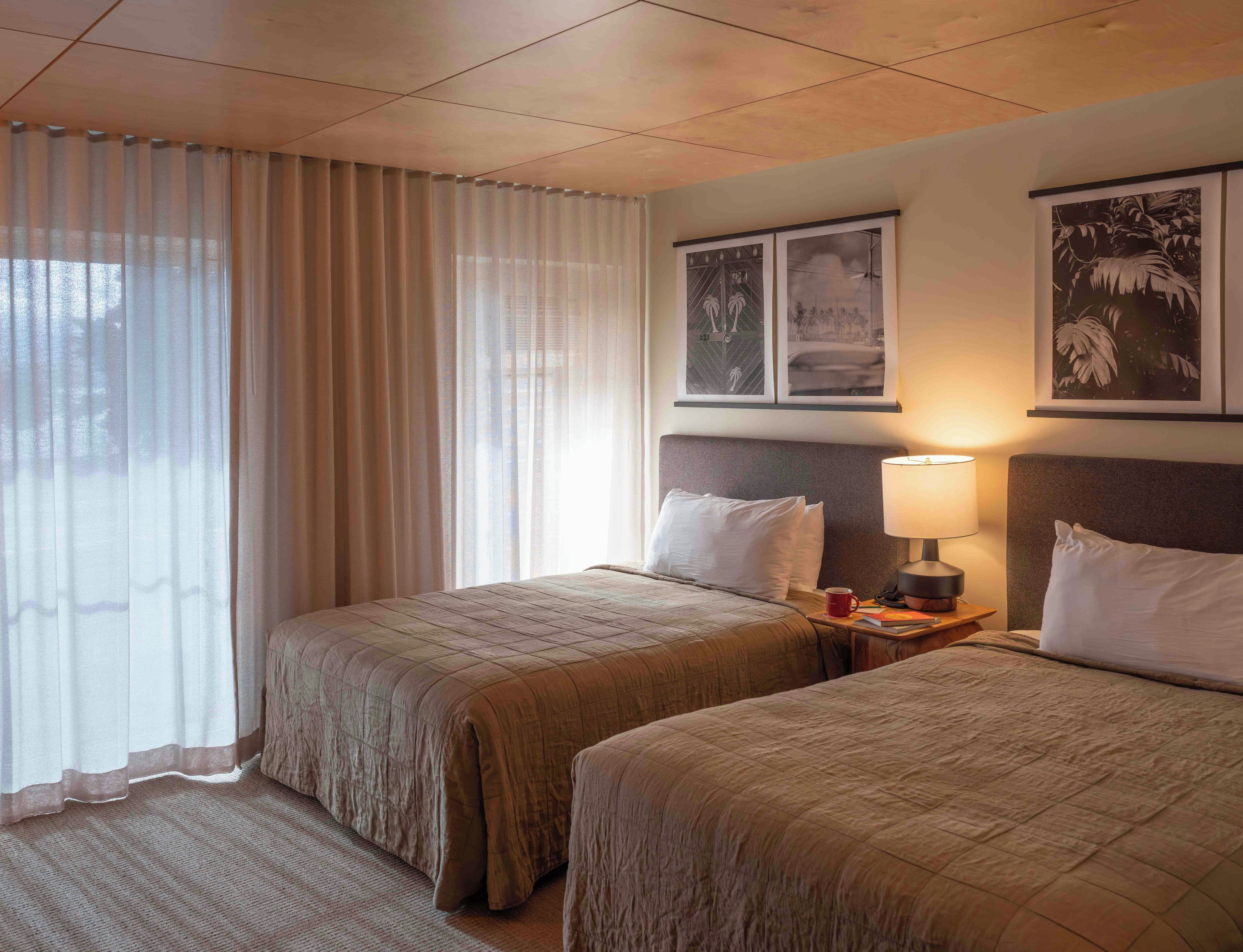 Habitación de hotel con dos camas, una ventana con cortinas translúcidas, una mesita de noche con lámpara y cuadros en la pared.
