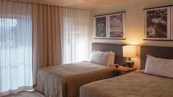 Habitación de hotel con dos camas, una ventana con cortinas translúcidas, una mesita de noche con lámpara y cuadros en la pared.