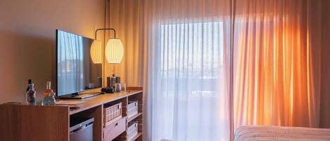 Habitación de hotel con cama, escritorio con computadora y lámpara, televisor y ventana con cortinas.
