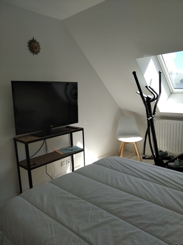 A bedroom with a bed, a TV on a shelf, a standing exercise bike, and a window with a view.