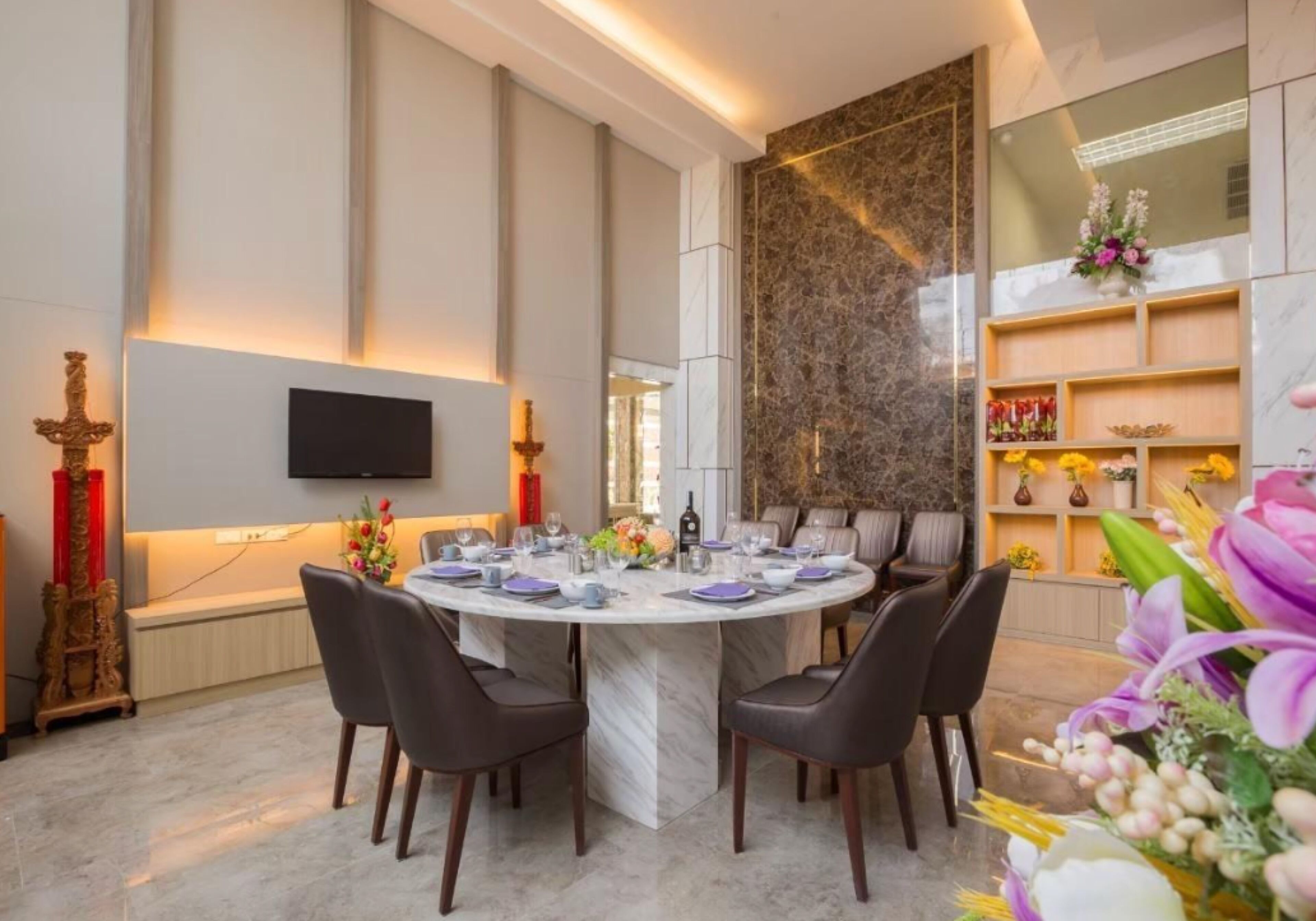 A dining area with a marble table, dark chairs, a flat-screen TV mounted on the wall, and a decorative wall with a floral arrangement.