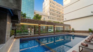 Una piscina con cerramiento de vidrio, sillas de descanso y un edificio con fachada de vidrio al fondo.