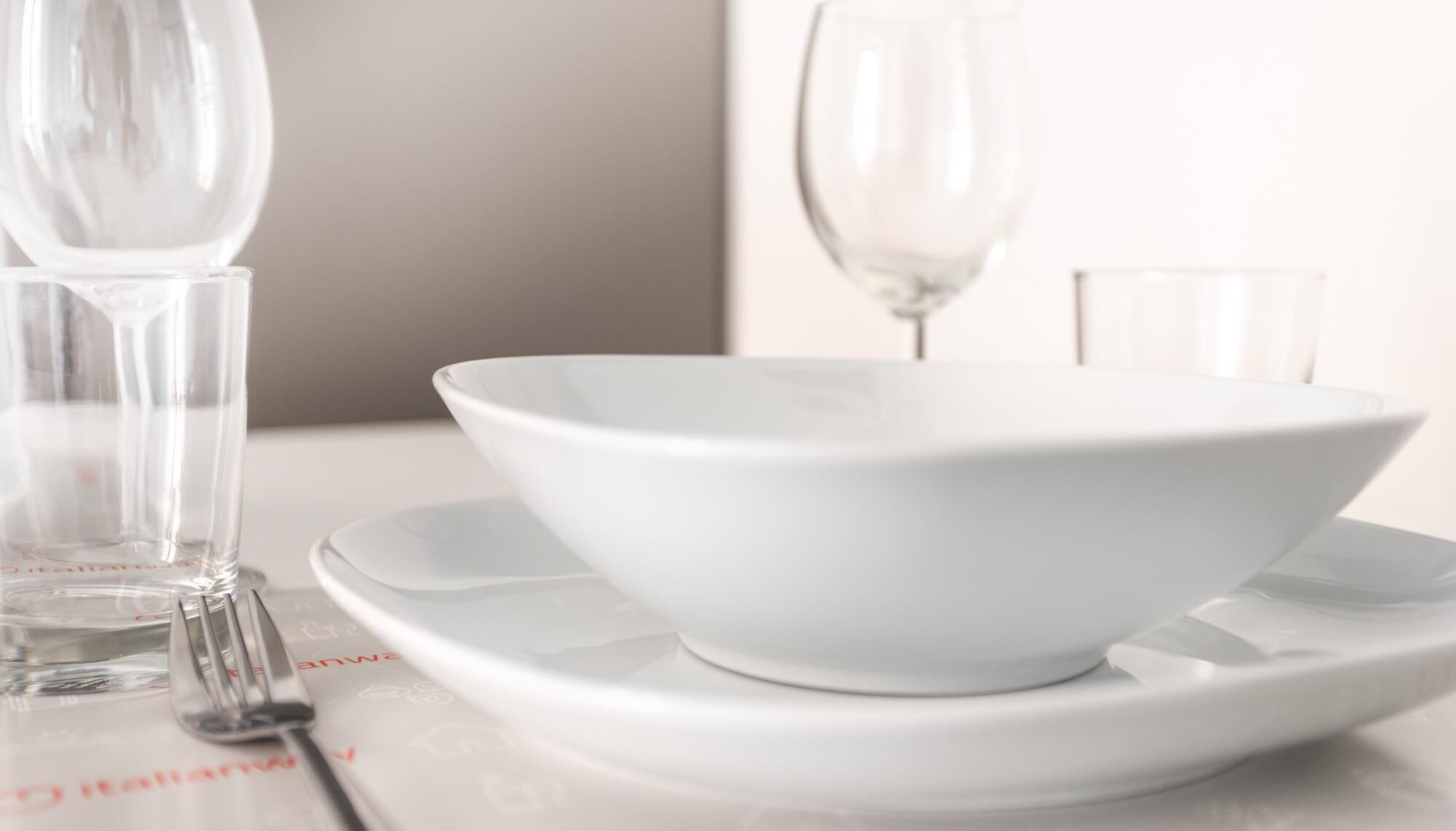 A white ceramic bowl on a white plate, accompanied by a fork and two clear glasses, likely for water or wine, on a table with a white tablecloth.