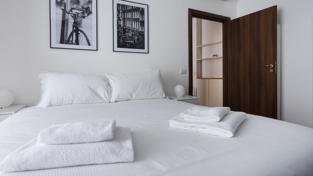 A neatly made bed with white linens and towels, a wooden wardrobe, and two black-and-white photographs on the wall. - Italianway Easy - Carlone 5 (Milan)