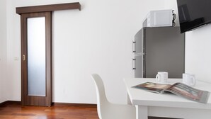 A room with a wooden door, a white desk with a magazine, a white chair, a microwave, and a television. - Italianway Easy - Carlone 5 (Milan)