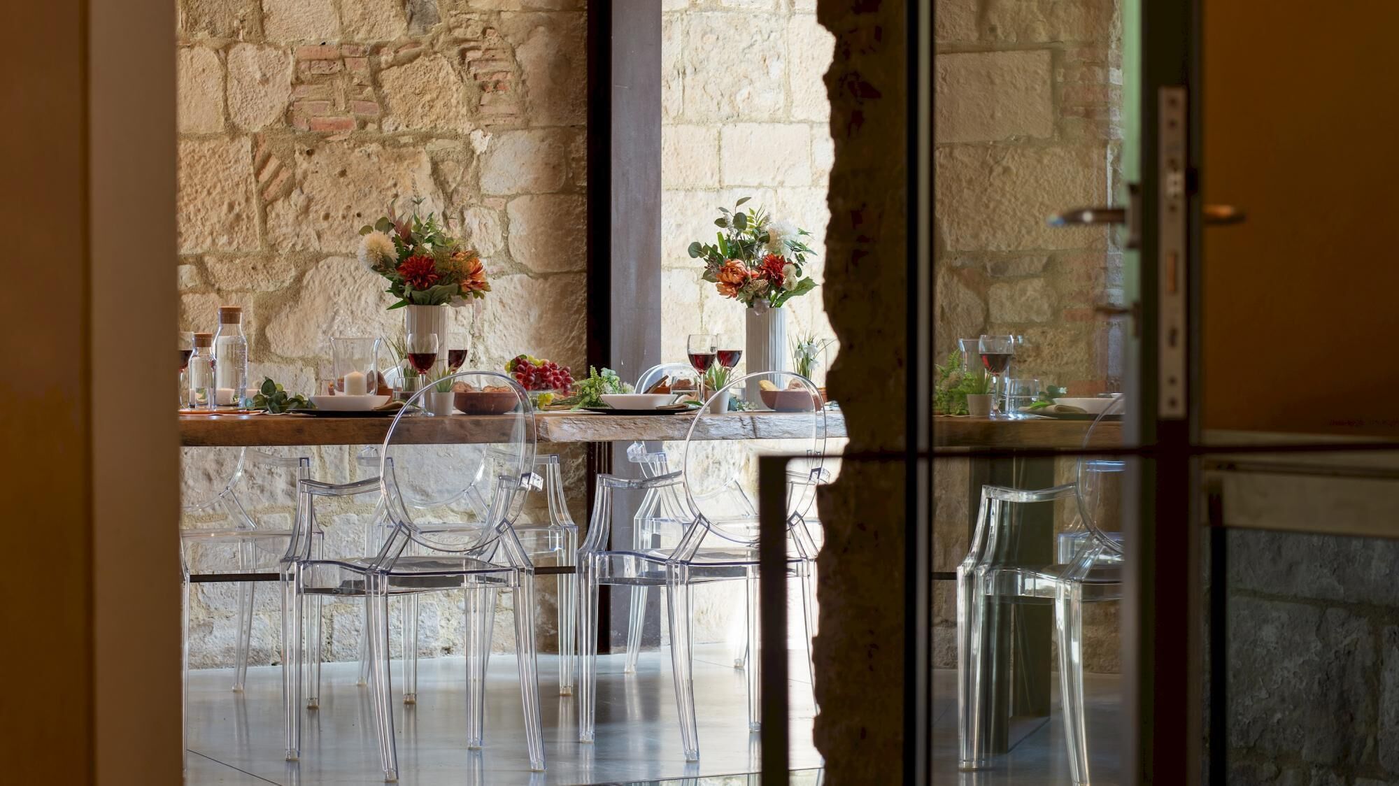 Un comedor con un fondo de pared de piedra, sillas de vidrio transparente, una mesa de madera dispuesta con platos, vasos y un florero.