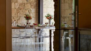 A dining area with a stone wall backdrop, clear glass chairs, a wooden table set with plates, glasses, and a vase of flowers. - Pieve di San Giovanni 10 (Pomarance)