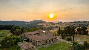 A stone building with a tiled roof, surrounded by greenery and hills in the background. - Pieve di San Giovanni 10 (Pomarance)