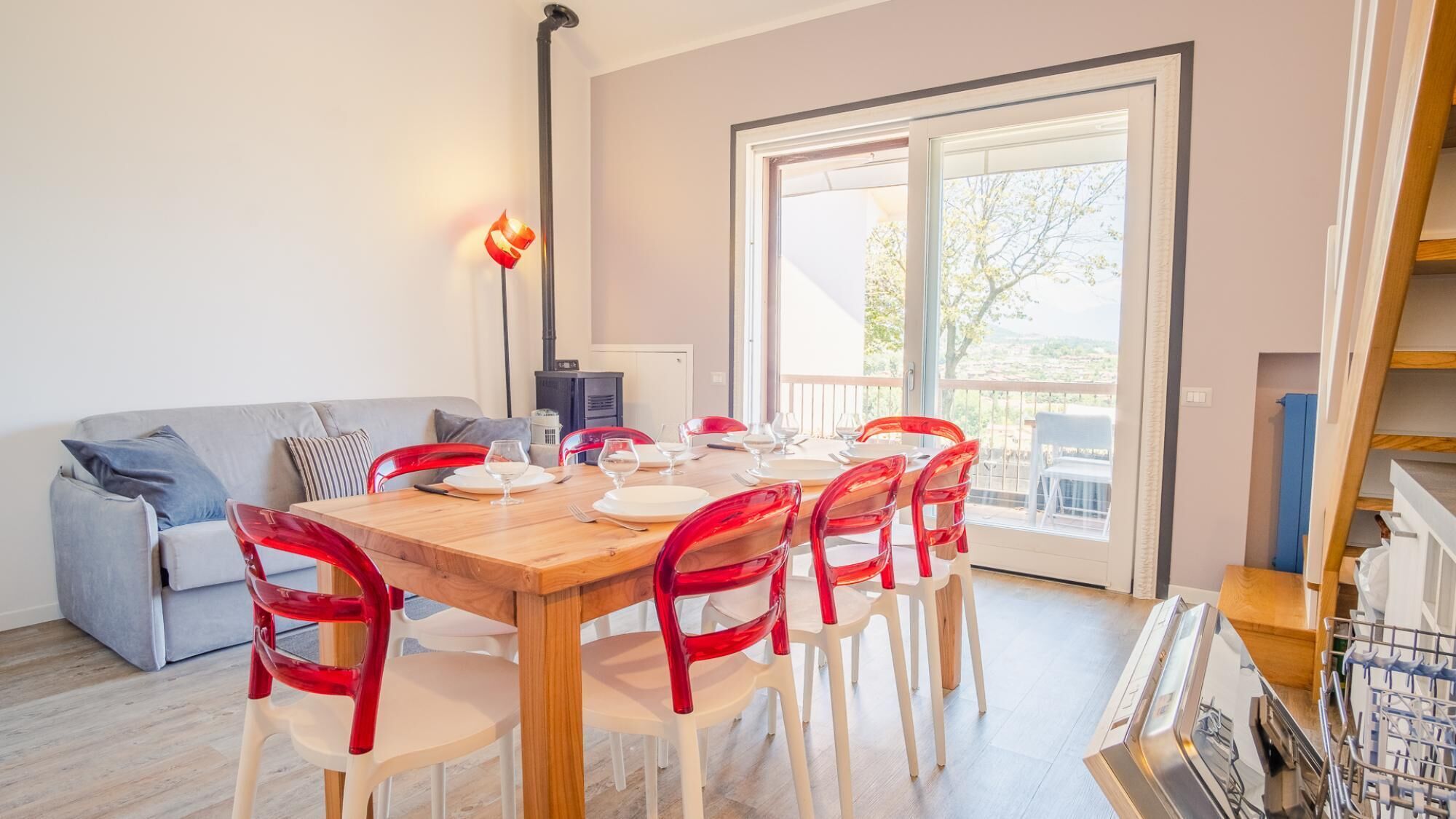 Een eethoek met een houten tafel gedekt voor vier personen, omringd door rode stoelen, een grijze bank en een open haard.