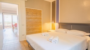 A hotel room with a bed, a wooden panel wall, a nightstand with a lamp, and a view of an outdoor balcony with red chairs.