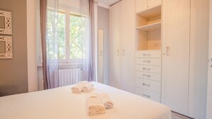 Een net opgemaakt bed met opgerolde handdoeken, een raam met dunne gordijnen en een witte kledingkast met meerdere lades.