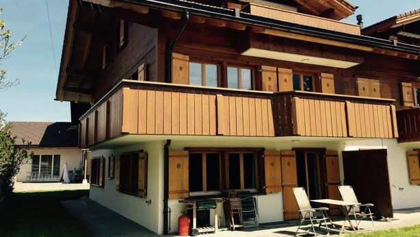 A two-story wooden house with a balcony, a small outdoor seating area, and a visible garage door. - Alpine Retreat by Interhome (Adelboden)