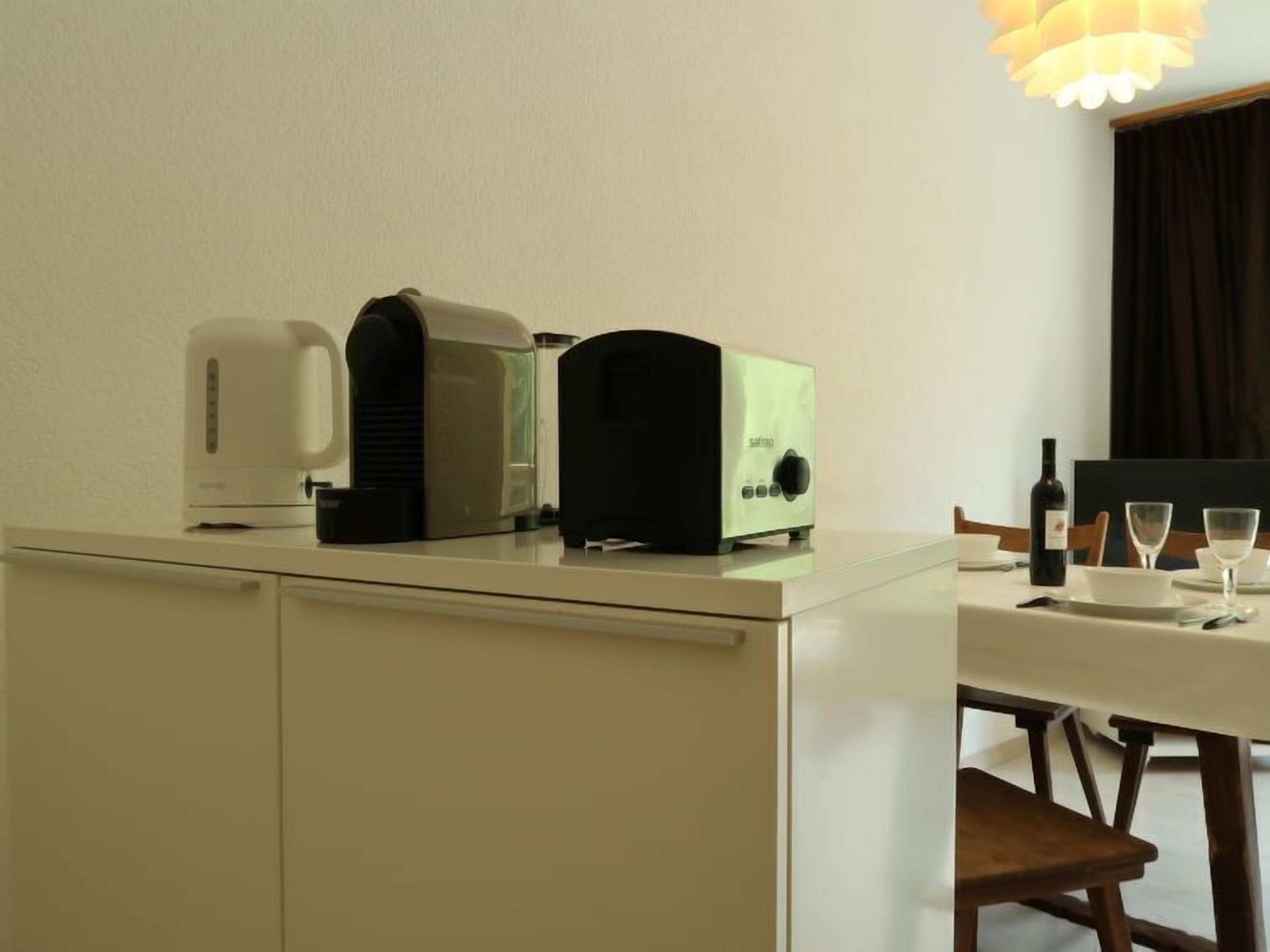 Une petite cuisine équipée d’un grille-pain, d’une machine à café et d’un four à micro-ondes. On y trouve également une table à manger avec des chaises et une bouteille de vin.