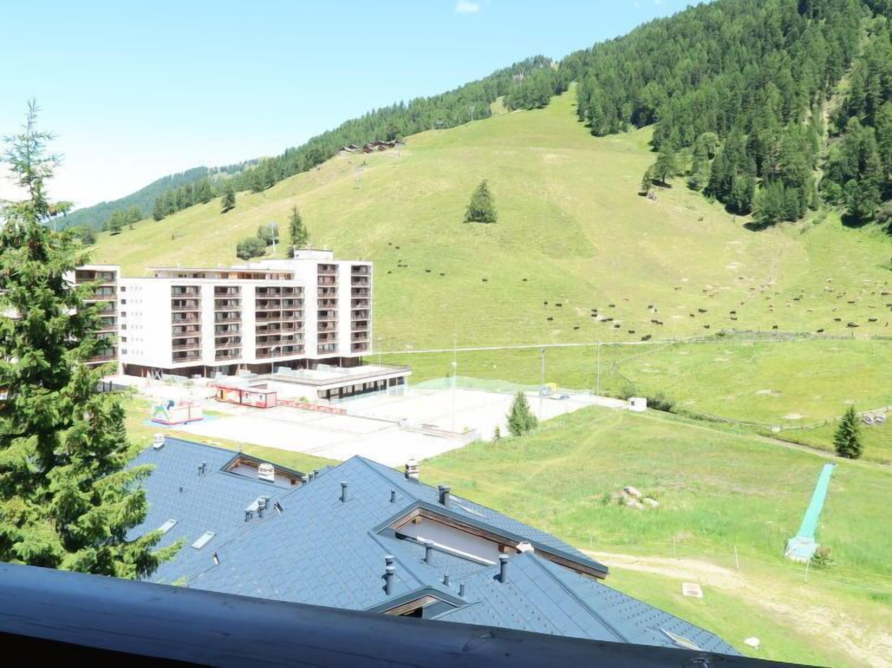 A hotel with multiple floors, a parking area, and a view of a green hillside with scattered trees and a few buildings.