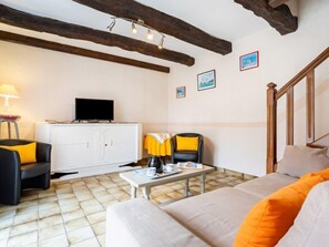 Une pièce chaleureuse avec un canapé, un fauteuil, une petite table sur laquelle se trouvent une tasse et une soucoupe, un téléviseur posé sur un meuble, et des poutres en bois au plafond.
