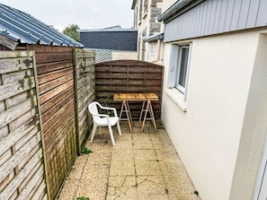 Un petit espace extérieur avec une table en bois et une chaise blanche, une clôture en bois et un bâtiment doté d’une fenêtre.