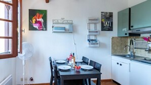 A compact kitchen with a dining table set for four, a white fan, and a window with blinds. - Les Cyprès by Interhome (Bandol)
