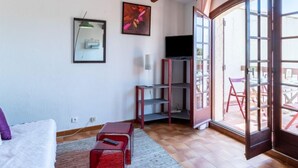 A living room with a sofa, a red coffee table, a TV on a stand, and a large window with an open door leading outside. - Les Cyprès by Interhome (Bandol)
