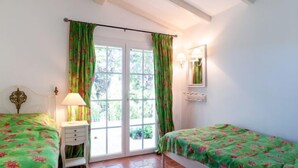 Une chambre avec deux lits, une table de chevet, une lampe, une fenêtre avec des rideaux, un miroir et un sol carrelé.