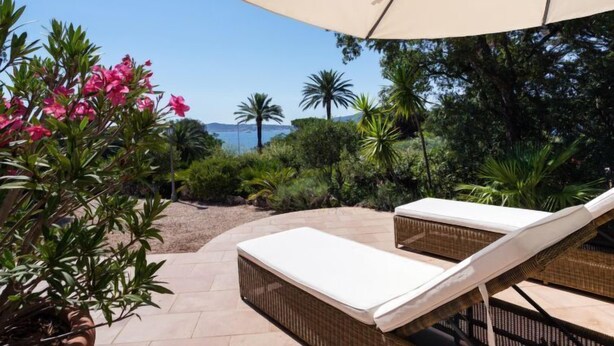 Une terrasse dotée de deux chaises longues et d’un grand parasol, entourée d’une végétation luxuriante et offrant une vue sur la mer.