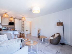 Un salon moderne avec un canapé, une table basse et un fauteuil en osier. On aperçoit à l’arrière-plan un coin cuisine avec une table à manger et des chaises.