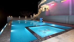 Una piscina con borde de mármol, una escalera que lleva a un nivel superior y un edificio con balcones e iluminación exterior.