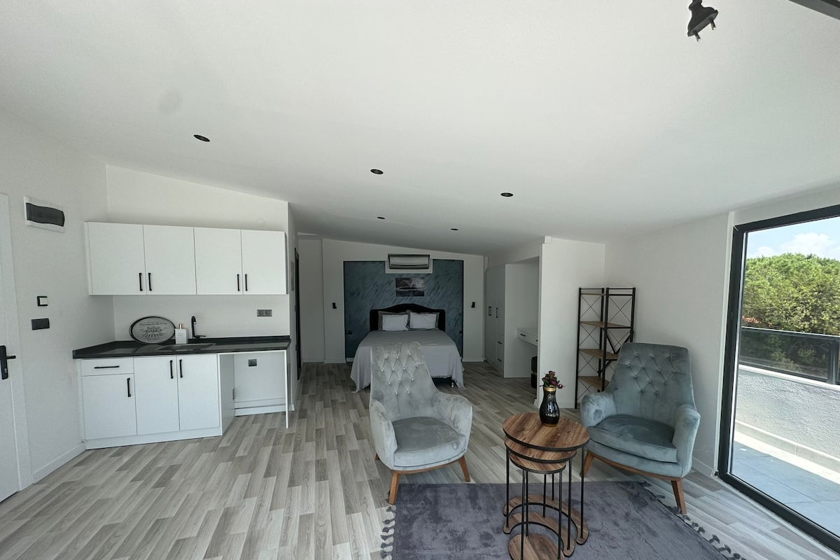 Un espacio habitable moderno y funcional con kitchenette, un dormitorio con cama grande, dos sillones y una pequeña mesa de centro.
