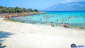 A crowded beach with clear turquoise water and mountains in the background.