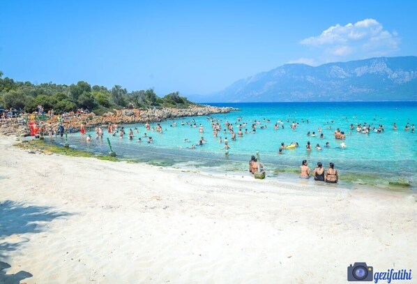 A crowded beach with clear turquoise water and mountains in the background. - Hotel Nil (Ula)