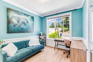 Une pièce avec un mur de couleur turquoise, un grand tableau abstrait, un canapé bleu avec un plaid blanc, un bureau en bois et une vue sur la verdure à l’extérieur.