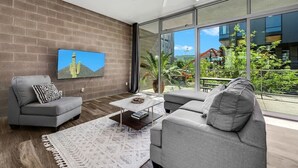 Un salon moderne avec une grande télévision, des canapés gris, une table basse et une vue sur un balcon agrémenté de plantes.