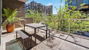 Un balcon avec une table et un banc, des plantes en pot et une vue sur des immeubles modernes.