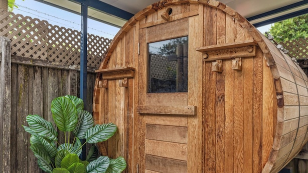 Une structure en forme de tonneau, faite de bois, avec une porte et une fenêtre, entourée d’une clôture en bois et agrémentée d’une plante en pot.