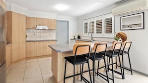 Une cuisine compacte avec des meubles en bois, un îlot central intégré doté de tabourets, une fenêtre avec stores et un climatiseur fixé au mur.
