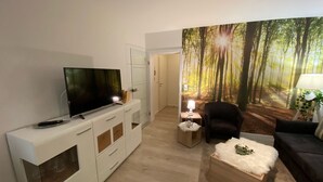 A modern living room with a flat-screen TV, a white entertainment unit, a cozy armchair, a small wooden coffee table, and a forest-themed wall mural. - Waldläufer Brilon (Brilon)